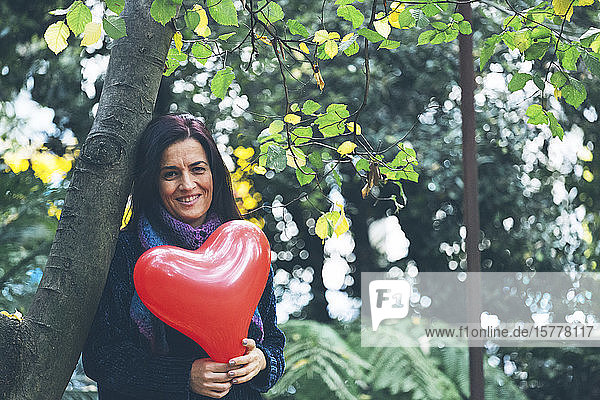 Lächelnde Frau hält Herz Ballon durch Bäume