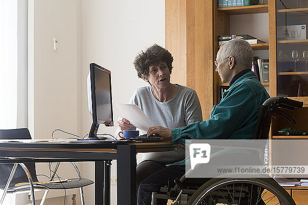 Senior woman sitting in a wheelchair at a computer  helper assisting with paperwork.