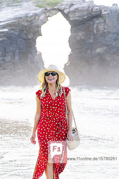 Blonde Frau in rotem Kleid und Hut  die am Strand spazieren geht  Natürlicher Bogen an der Playa de Las Catedrales  Spanien