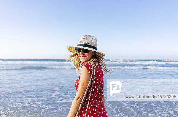 Blonde Frau in rotem Kleid und Hut am Strand  die sich dreht und in die Kamera schaut