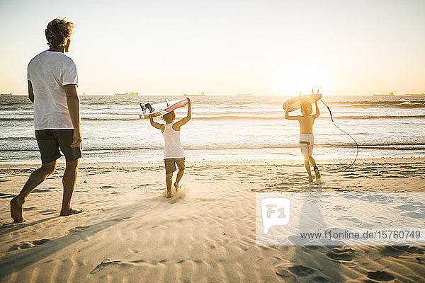 Vater und zwei Söhne mit Surfbrettern bei Sonnenuntergang am Strand spazieren