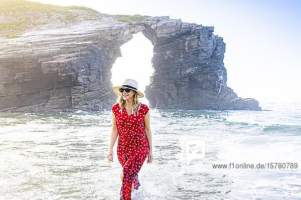 Blonde Frau in rotem Kleid und Hut die am Strand spazieren geht Natürlicher Bogen an der Playa de Las Catedrales Spanien