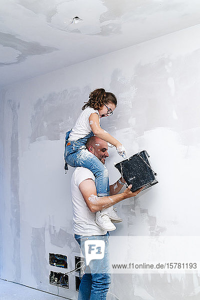 Mädchen sitzt auf den Schultern des Vaters und streicht die Wand ihres neuen Hauses