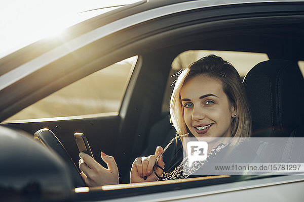 Portrait of smiling young woman with smartphone sitting in her car in the evening