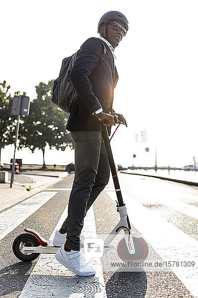 Businessman with backpack and cycling helmet crossing street on push scooter at sunset