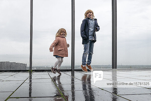 Brother and little sister jumping on view terrace on rainy day