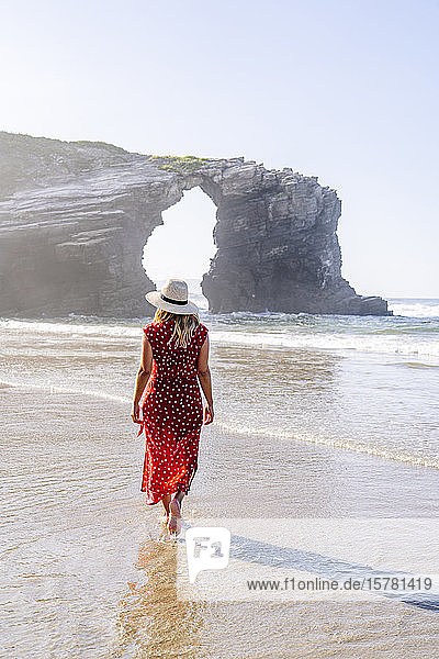 Blonde Frau in rotem Kleid und Hut  die am Strand spazieren geht  Natürlicher Bogen an der Playa de Las Catedrales  Spanien