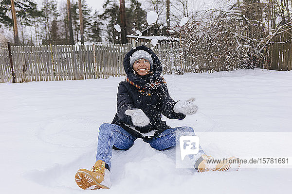 Happy woman sitting on snow field playing with snowballs