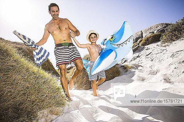 Father and son with inflatable shark walking through the dunes to the beach