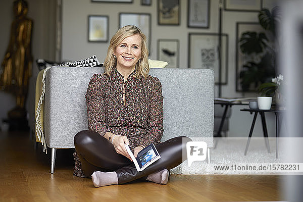 Porträt einer lächelnden blonden Frau  die zu Hause auf dem Boden sitzend entspannt