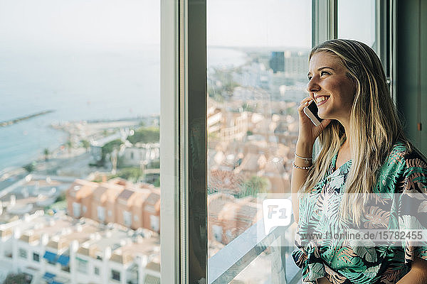 Lächelnde Frau am Telefon am Fenster in Küstenstadt