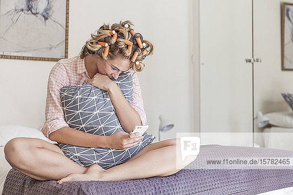 Glückliche junge Frau mit Lockenwicklern im Haar sitzt auf dem Bett und schaut auf ihr Handy