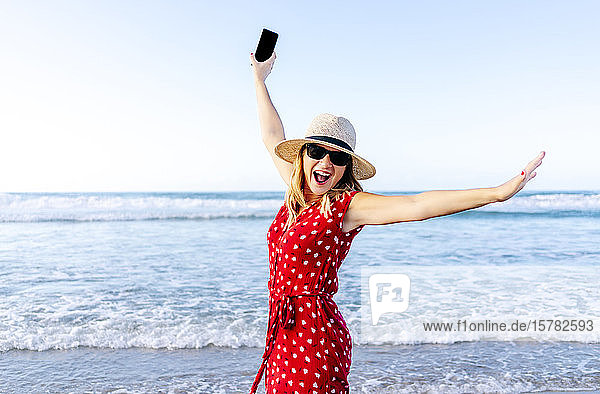 Lächelnde blonde Frau in rotem Kleid und Hut mit Smartphone in der Hand am Strand
