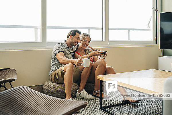 Glückliches Paar sitzt zu Hause am Fenster mit Kaffeetassen und Handy