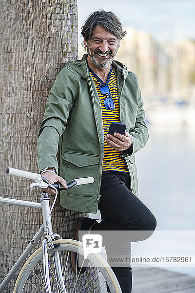 Porträt eines lachenden  reifen Mannes mit einem Fixie-Rad  das an einen Palmenstamm gelehnt ist  Alicante  Spanien