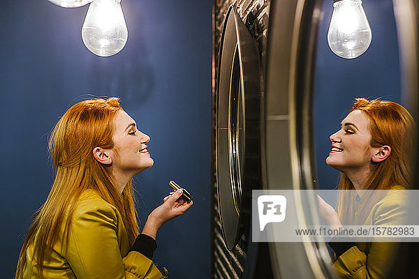 Lächelnde rothaarige junge Frau  die ihr Spiegelbild betrachtet und Lippenstift aufträgt