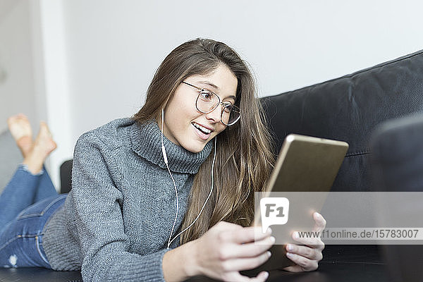 Porträt einer lächelnden jungen Frau  die mit Kopfhörer und digitalem Tablett auf der Couch liegt