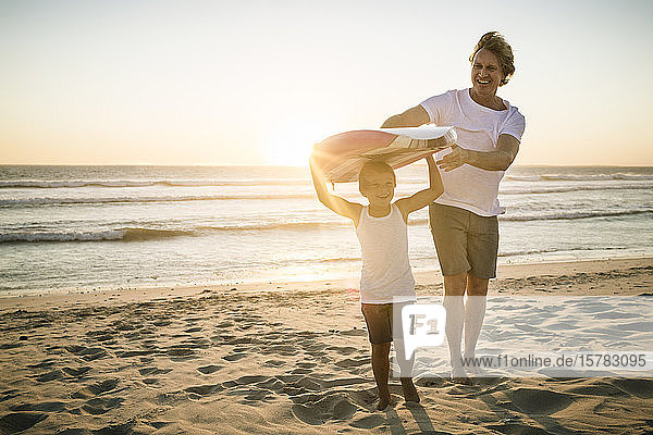 Happy father and son with surfboard walking on the beach at sunset
