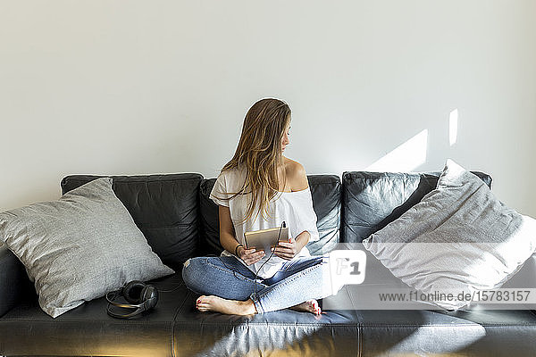 Junge Frau sitzt auf Couch mit Kopfhörern und digitalem Tablet
