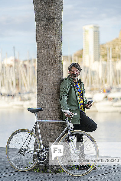 Porträt eines entspannten reifen Mannes mit einem Fixie-Rad  das an einen Palmenstamm gelehnt ist  Alicante  Spanien