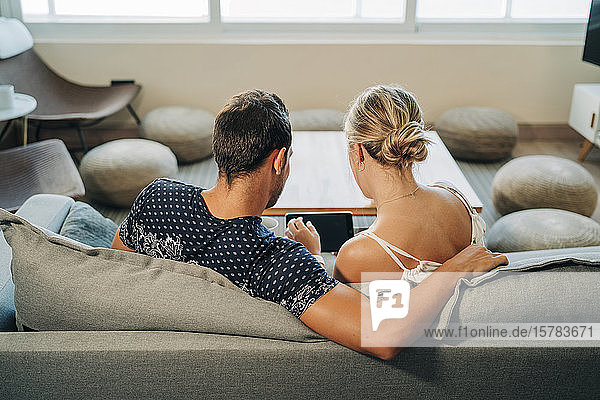 Rear view of couple sitting on couch in living room sharing a tablet