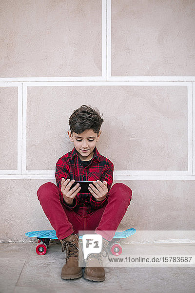 Junge sitzt mit einem Smartphone auf einem Skateboard an einer Wand