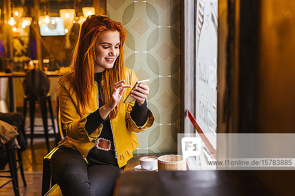 Porträt einer glücklichen rothaarigen jungen Frau  die mit ihrem Handy in einem Café sitzt