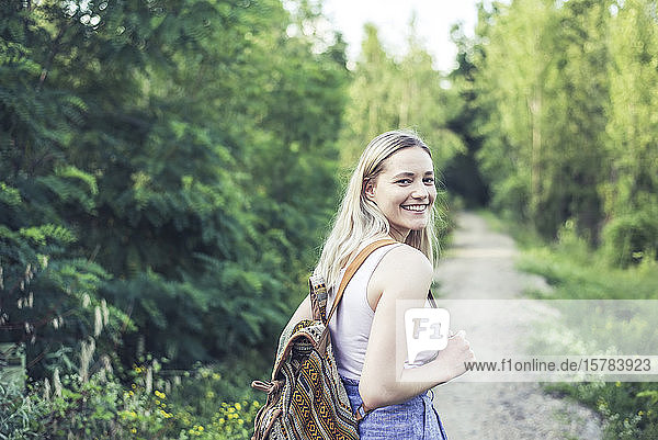 Porträt einer lächelnden jungen Frau mit Rucksack auf einem Waldweg