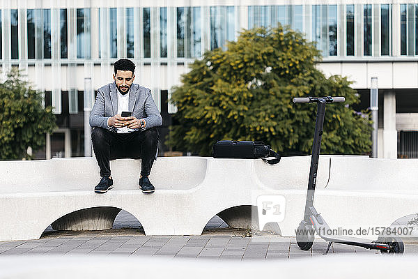 Casual young businessman sitting on a bench in the city using cell phone