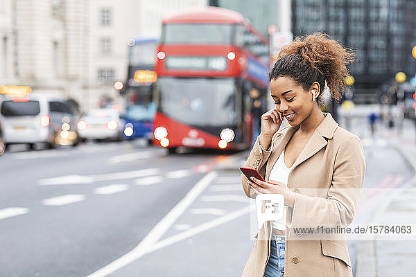 Lächelnde junge Frau mit Handy und Ohrstöpseln in der Stadt  London  Großbritannien