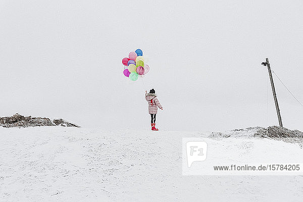 Rückenansicht eines Mädchens mit Ballons  das im Winter auf einem schneebedeckten Hügel steht