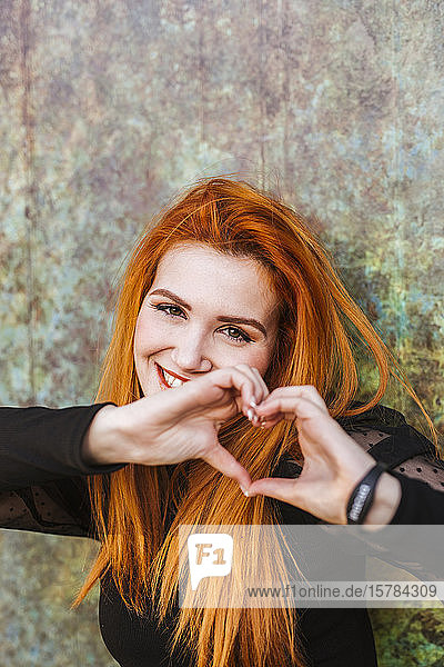 Porträt einer glücklichen rothaarigen jungen Frau  die mit ihren Fingern das Herz formt