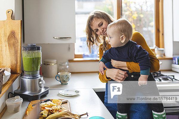 Woman in the kitchen preparing healthy smoothie and holding her baby son