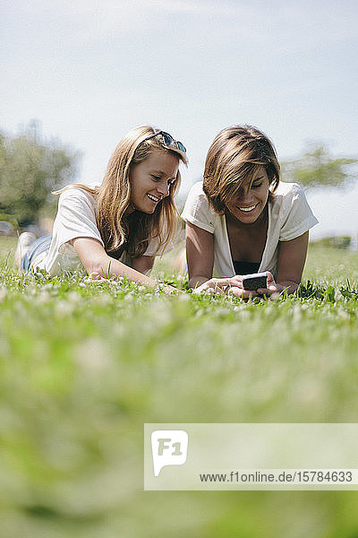 Zwei lächelnde Freundinnen liegen auf einer Wiese und teilen sich ein Handy
