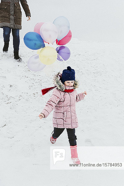 Kleines Mädchen mit Ballons läuft im Winter einen Hügel hinunter