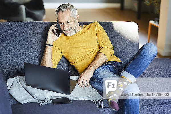 Mature man using cell phone and laptop on couch at home