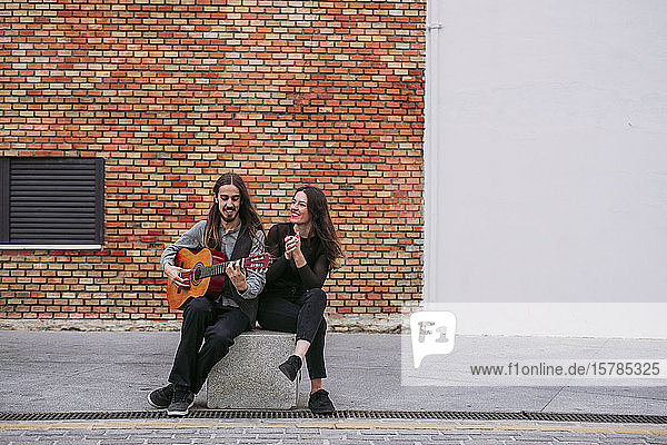 Zwei lächelnde junge Musiker sitzen auf einem Stein und spielen Gitarre