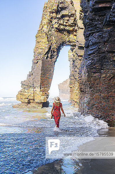 Blonde Frau in rotem Kleid und Hut die am Strand spazieren geht Natürlicher Bogen an der Playa de Las Catedrales Spanien