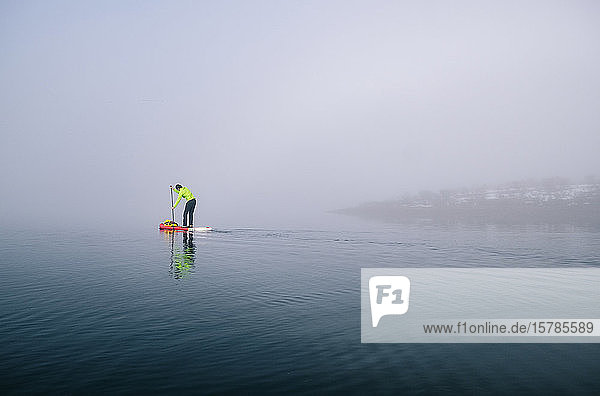 Mann steht beim Paddel-Surfen auf einem See im Nebel auf