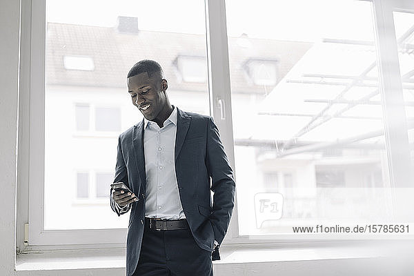 Portrait of smiling young businessman standing in front of window looking at cell phone