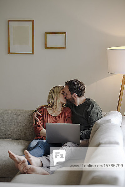 Paar sitzt zu Hause auf der Couch  küsst sich  benutzt einen Laptop