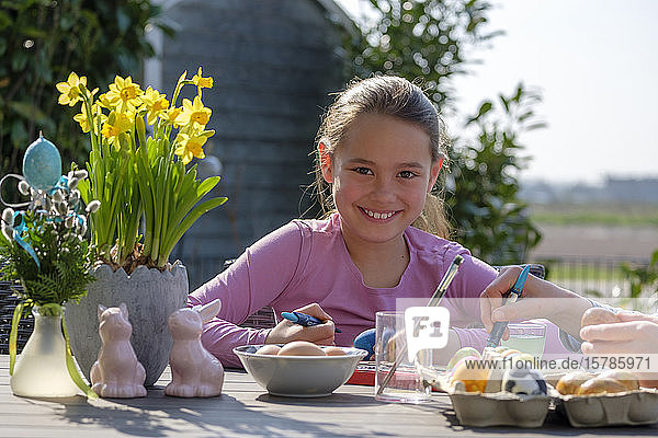 Porträt eines glücklichen Mädchens  das am Gartentisch sitzt und Ostereier malt