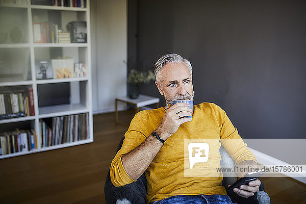 Entspannter reifer Mann zu Hause mit Handy und Tasse Kaffee