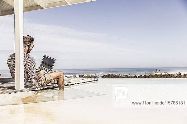 Man working on laptop next to pool at a beach house