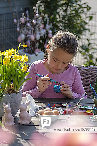 Konzentriertes Mädchen sitzt am Gartentisch und malt Ostereier