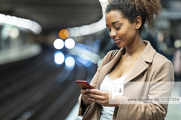 Lächelnde junge Frau benutzt Mobiltelefon am Bahnsteig der U-Bahn-Station