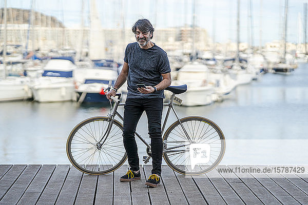 Portrait of smiling mature man with fixie bike and cell phone standing on boardwalk  Alicante  Spain