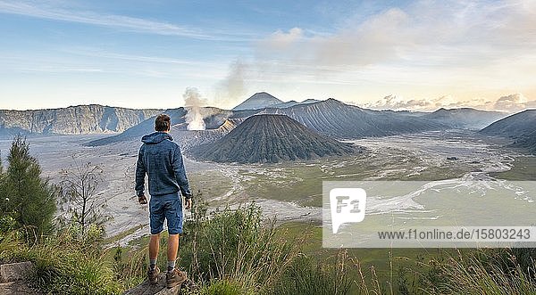 Junger Mann vor Vulkanlandschaft  Blick in Tengger Caldera  rauchender Vulkan Gunung Bromo  davor Mt. Batok  dahinter Mt. Kursi  Mt. Gunung Semeru  National Park Bromo-Tengger-Semeru  Java  Indonesien  Asien