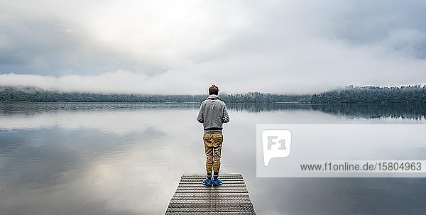 Young man standing on a jetty looking over a lake  foggy atmosphere  Lake Mapourika  West Coast  South Island  New Zealand  Oceania