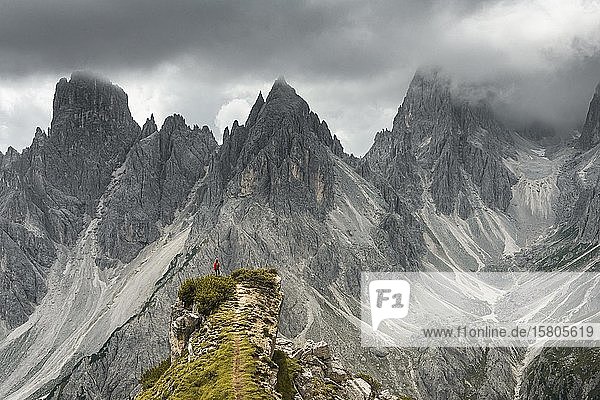 Mann mit roter Jacke steht auf einer Stufe  hinter ihm Berggipfel und spitze Felsen  dramatische Wolken  Cimon die Croda Liscia und Cadini Gruppe  Auronzo di Cadore  Belluno  Italien  Europa